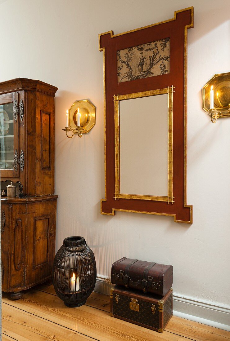 Nostalgischer Truhenstapel und Windlicht auf Holzboden vor Wandspiegel mit vergoldetem und bemaltem Rahmen, seitlich Wandleuchter aus Messing