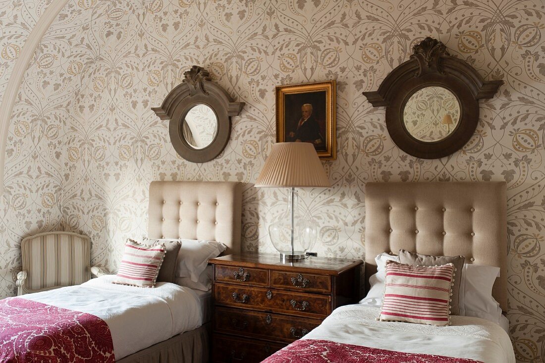 Zweibettzimmer mit floraler Tapete im Dachraum eines englischen Herrenhauses, Kopfteile mit Knopfpolsterung, antike Kommode und runde Spiegel mit Holzrahmen