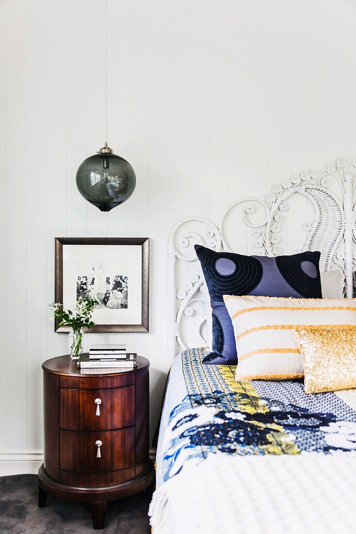 Cushions and throw on bed with ornate headboard next to spherical glass lamp above round bedside cabinet