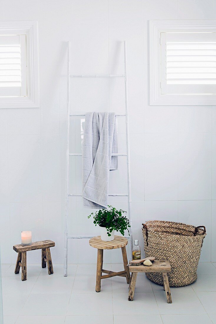 Still-life arrangement of ladder-style towel rail, wooden stools and basket in white bathroom
