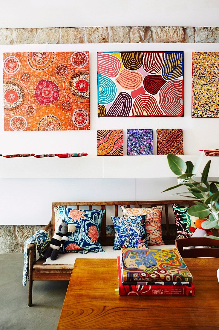 A gallery of colorful pictures over a wooden sofa bench with colorful pillows