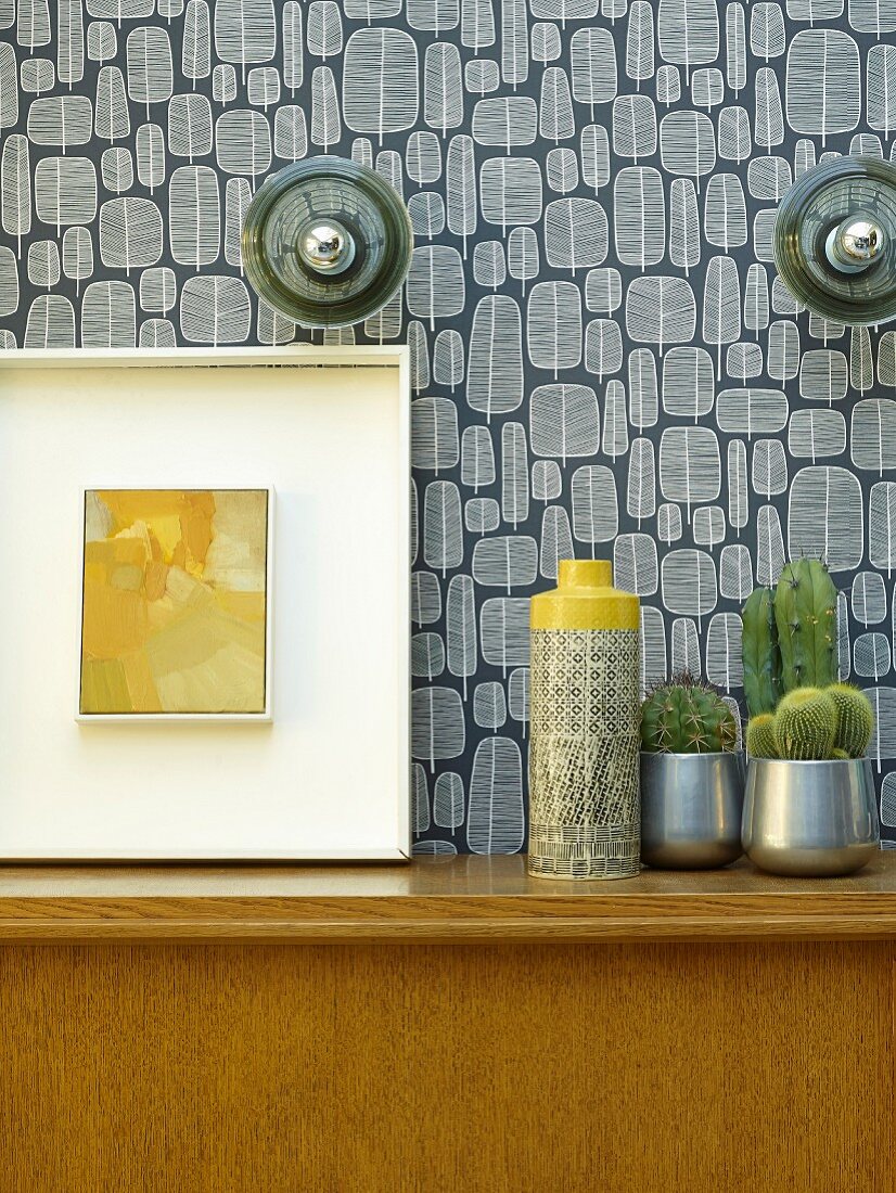 Vase neben Kakteen und Bild auf Holzschrank vor tapezierter Wand mit Retro Muster