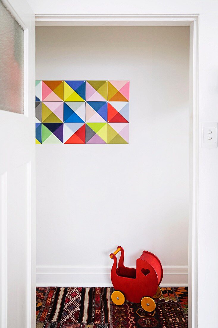 Red wooden retro dolls' pram on ethnic rug below geometric picture on wall
