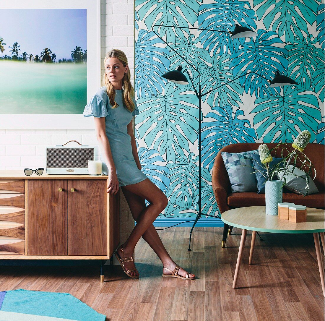 Frau an Sideboard in Retro Wohnraum mit braunem Ledersofa und Klassikerleuchte vor Tapete mit Blattmotiv