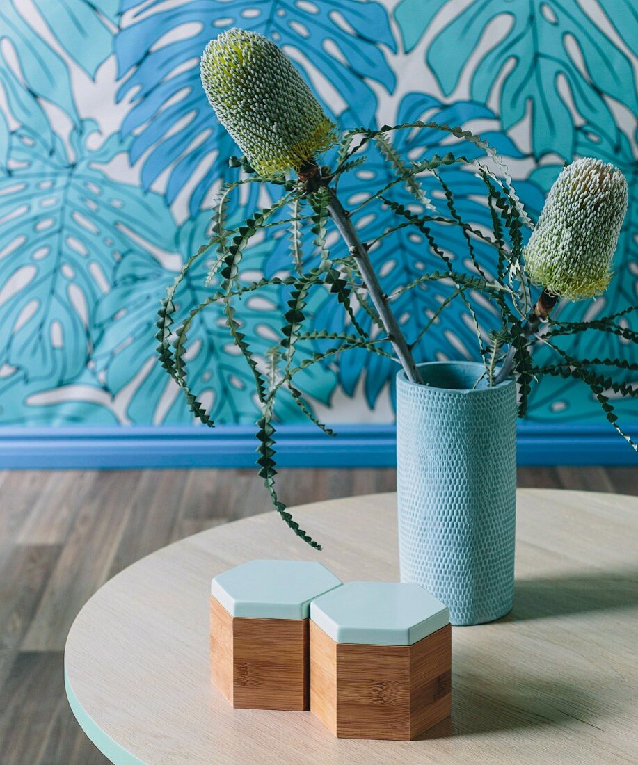 Exotische Blütenpflanze in hellblauer Base auf Tisch, dahinter Tapete mit Blattmotiv