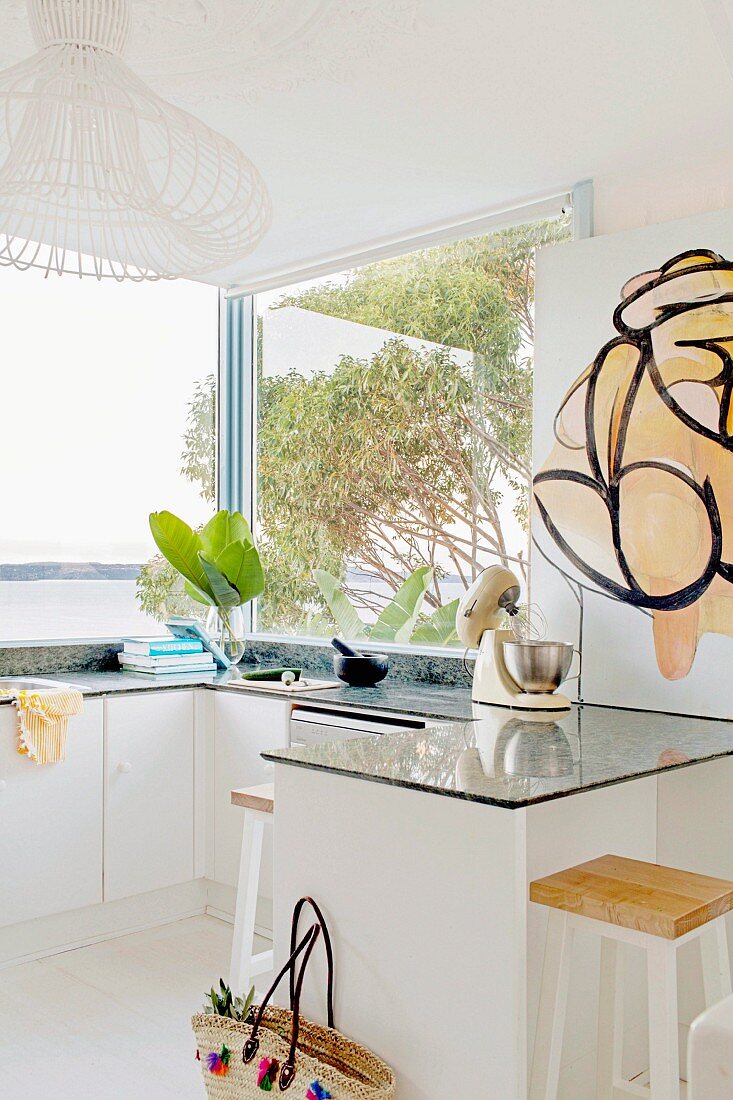 Modern, white kitchen with stone worktop in front of a window with a view