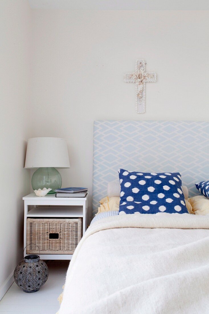 Bed with upholstered headboard and blue and white polka dot pillow, simple bedside table with drawer and cross on the wall
