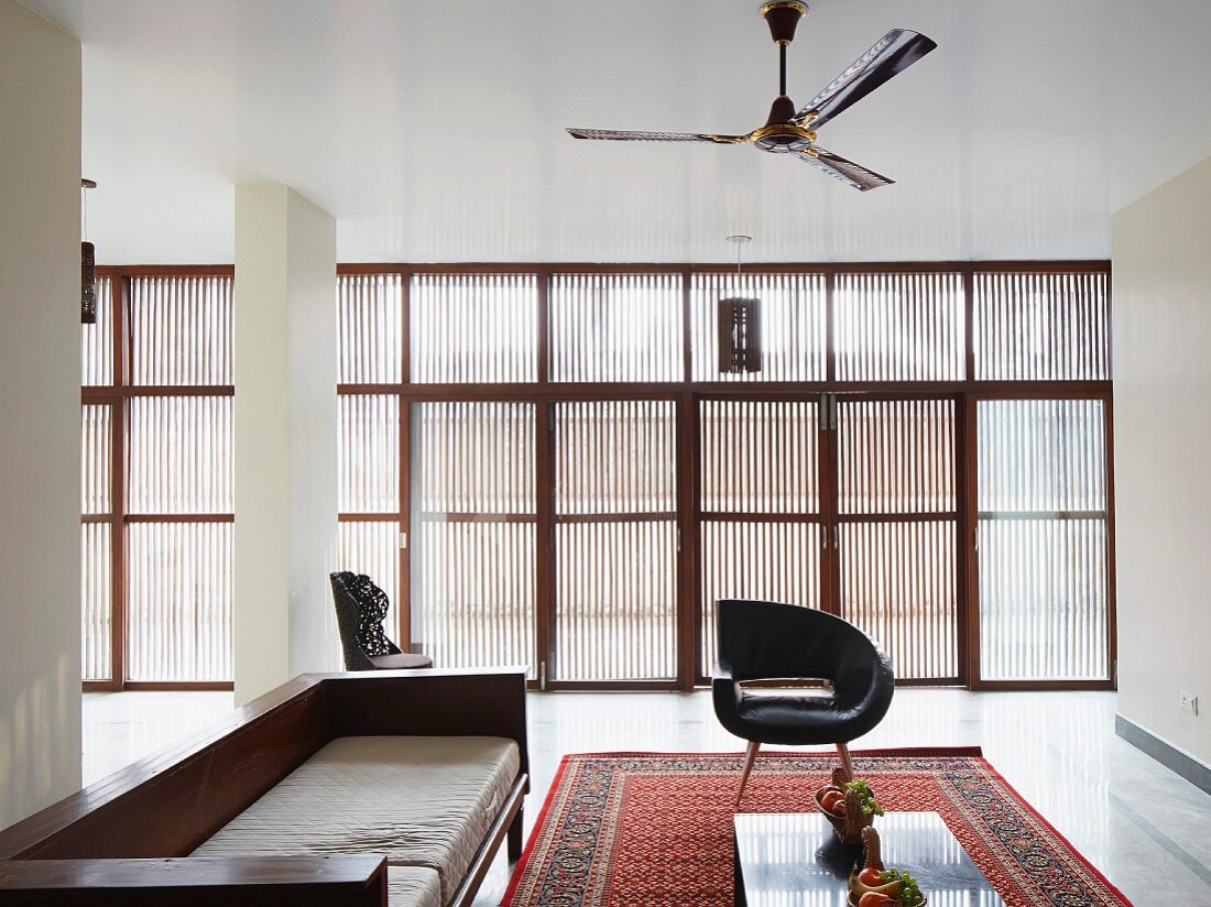 Seating area with retro leather armchairs and purist wooden sofa on Oriental rug; subdued light falling through glass wall with vertical, slatted sun screens
