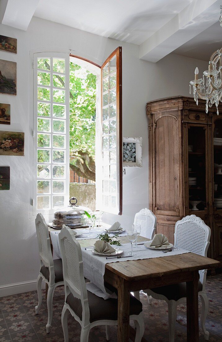 Rokoko Stühle um gedeckten Tisch vor offenem Fenster, seitlich alter Bauernschrank in ländlichem Esszimmer