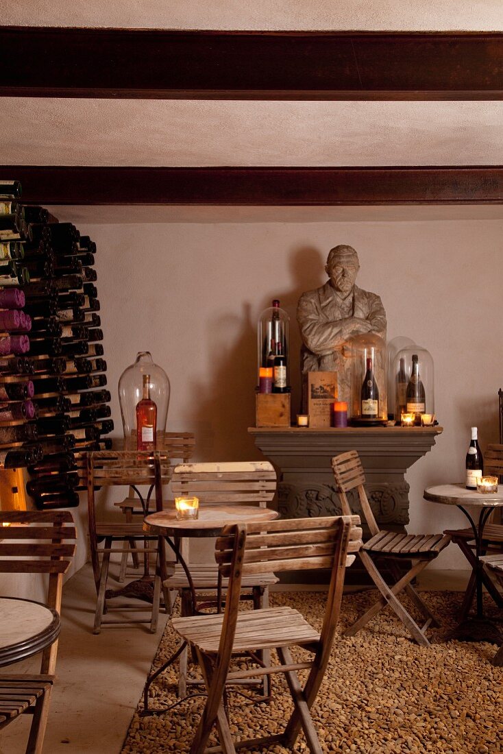 Folding wooden chairs around bistro table in front of stone bust on antique console table and wine rack in cellar-style room