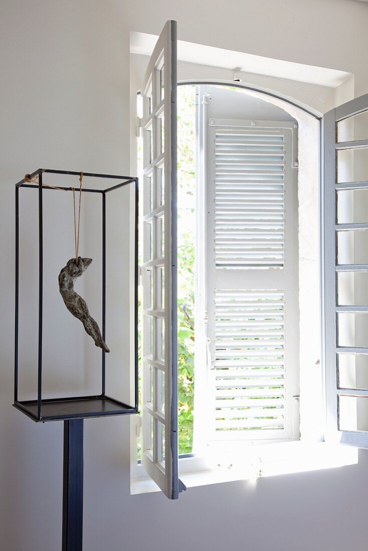 Suspended figurine in glass case on metal stand next to lattice window with white interior shutters