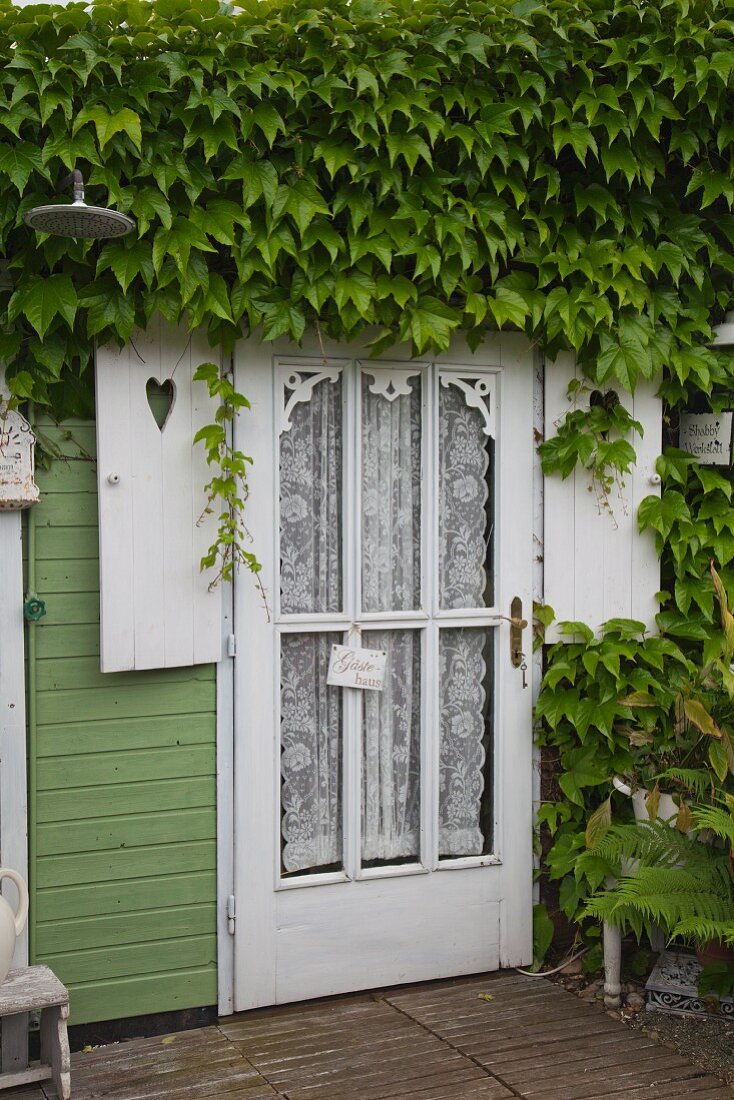 Door of wooden house with lace curtains behind glass panels and climber-covered façade