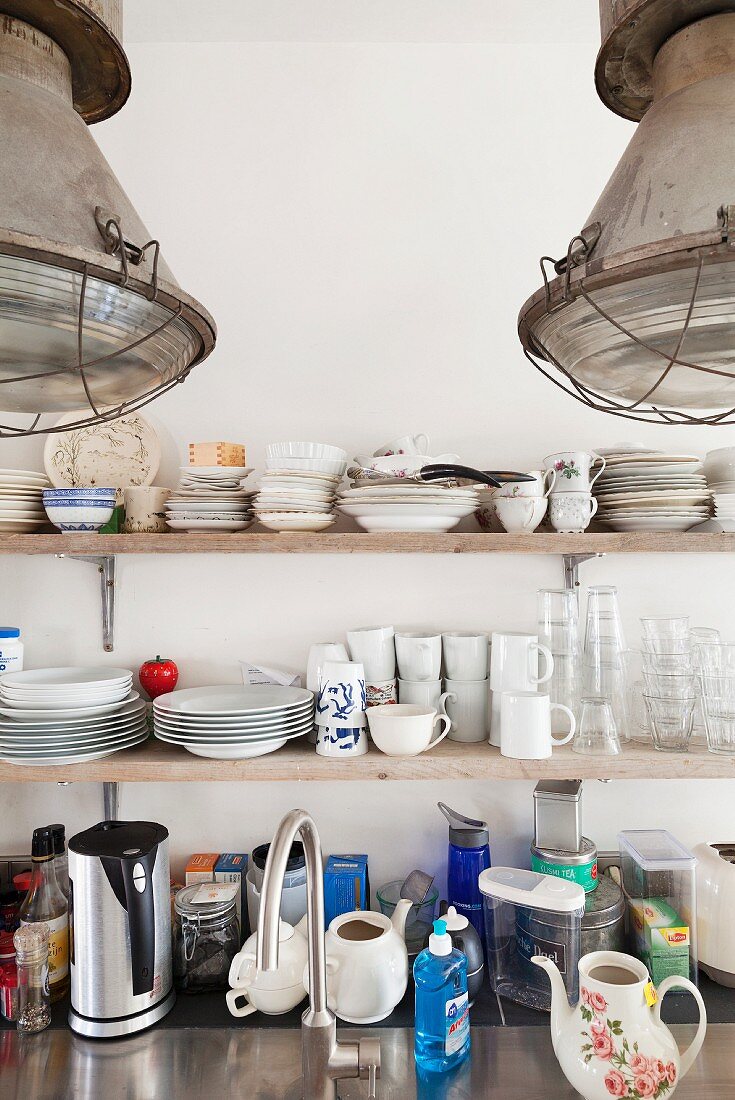 Detail of kitchen; crockery on wooden shelves, utensils on stainless steel worksurface and industrial-style pendant lamps