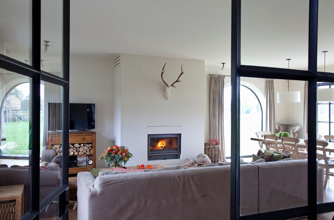 View through glass wall into living room with dining table and fire in fireplace