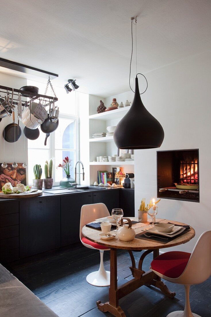 Essplatz mit weißen Klassikerstühlen unter schwarzer Pendelleuchte vor Küchenzeile in eklektischem Ambiente mit offenem Kaminfeuer und Grilleinsatz