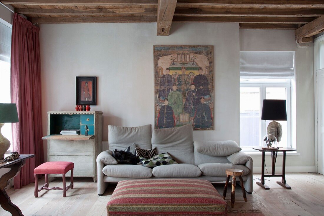 Ottoman and pale grey sofa in rustic interior with ethnic artworks