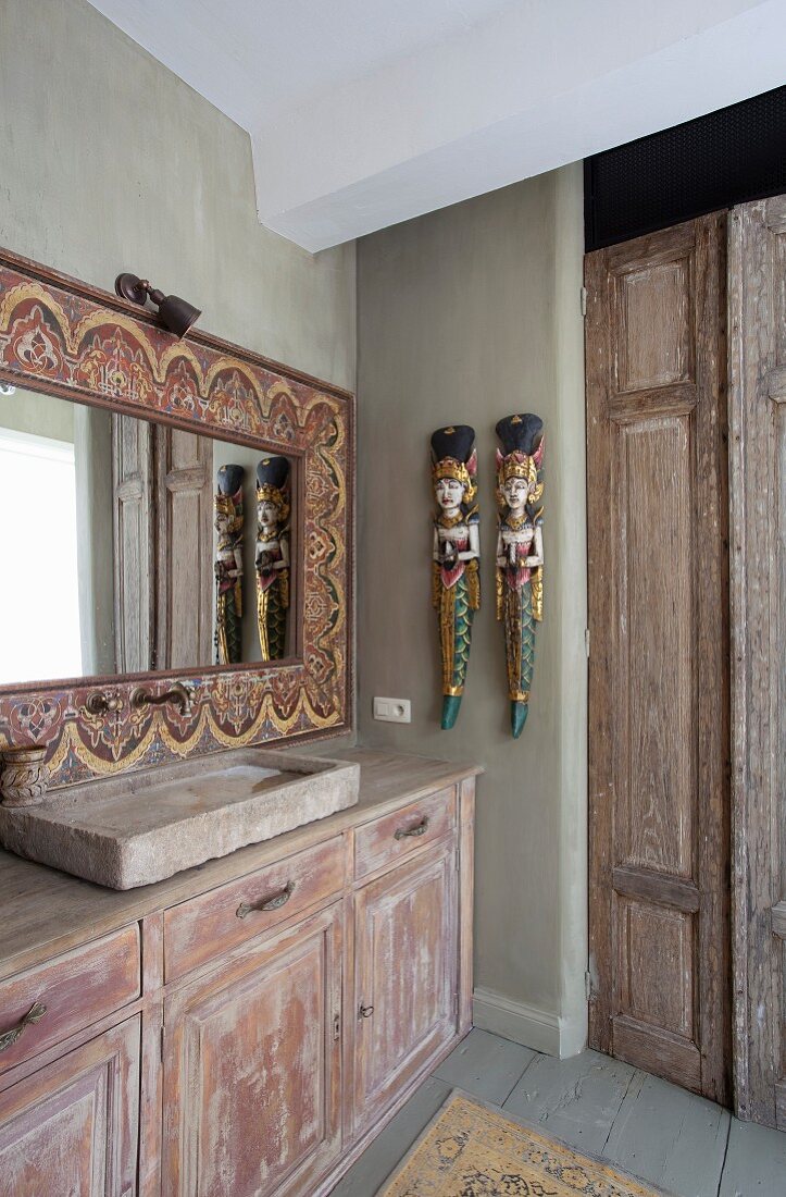 Vintage-Waschtisch mit Unterschrank an Wand, Ethno-Spiegel mit verziertem Holzrahmen und bemalte, asiatische Skulpturen an Wand