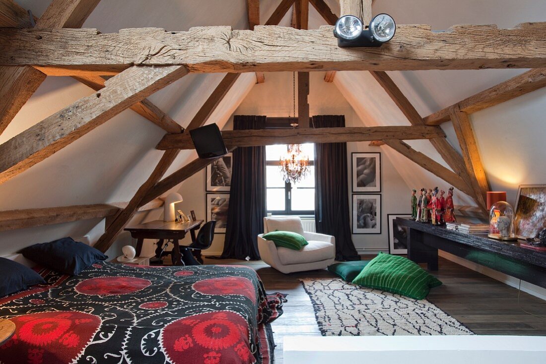 Bett mit gemusterter Tagesdecke in Dachraum mit sichtbarer, alter Holzbalkenkonstruktion