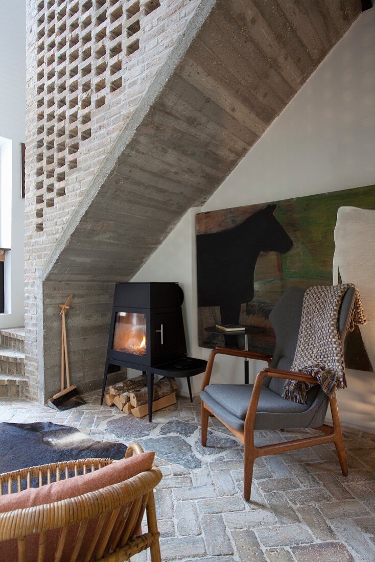 50s-style chair on herringbone stone floor and log burner below concrete staircase with perforated brick side wall
