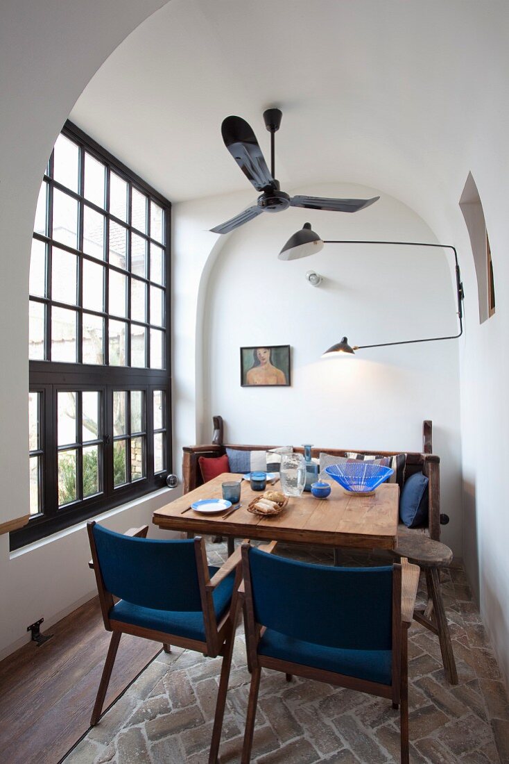 Blue upholstered chairs around wooden table below ceiling fan and retro wall lamps in dining room with lattice window