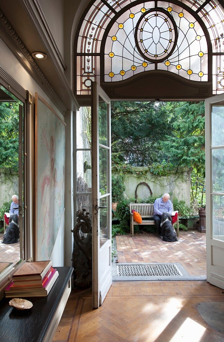 Offene Glasflügeltür mit Jugendstil Oberlicht und Blick in sommerlichen Innenhof mit älterem Herrn und Hund