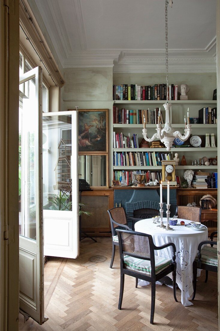 Traditioneller Salon; Armlehnstühle um runden Tisch unter Kronleuchter, im Hintergrund Bücherwand und romantisches Gemälde