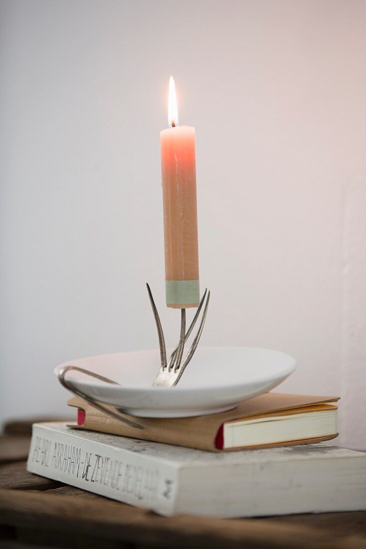 Brennende Kerze auf Kerzenhalter aus gebogener Vintage-Gabel mit Untertasse auf Bücherstapel