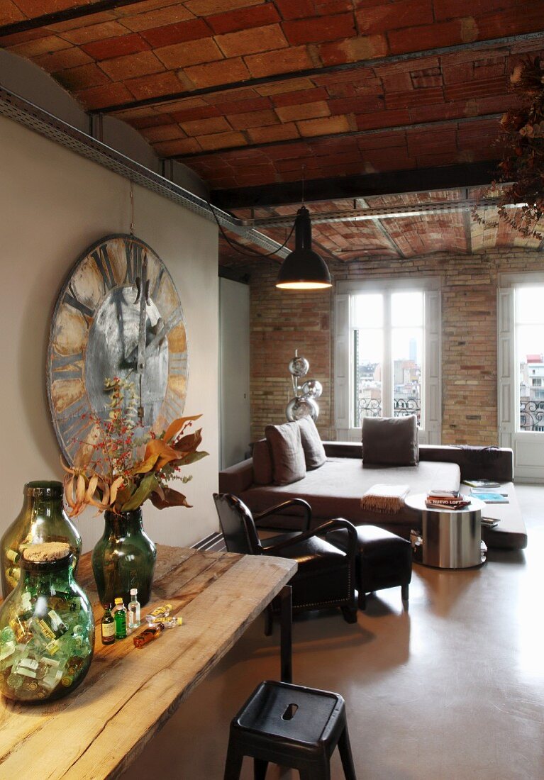 Vasengruppe auf Holztisch, davor Metallhocker im Retro Stil, im Hintergrund Wohn-, Schlafbereich in rustikalem Ambiente mit Wand und Decke aus Ziegel