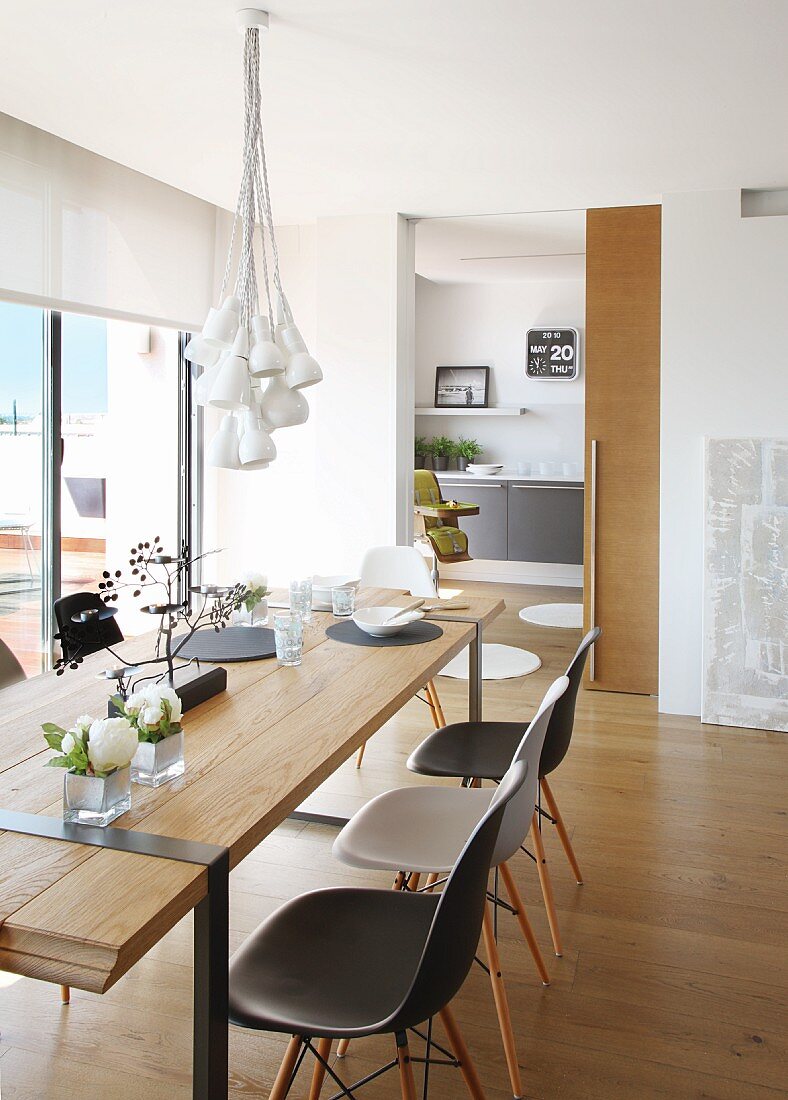Leuchtenbündel über schlichtem Designerholztisch mit Eames Plastic Chairs; im Hintergrund eine Schiebetür zur Küche