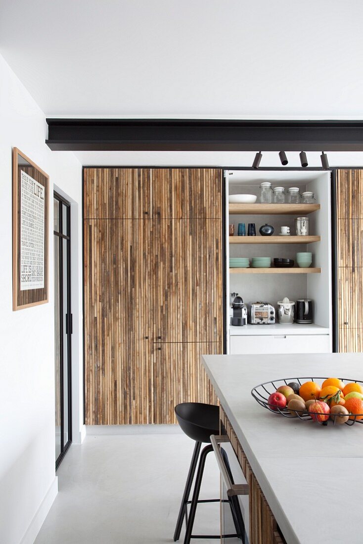 Kitchen counter in front of fitted cupboards with slatted wooden fronts and open-fronted shelving element
