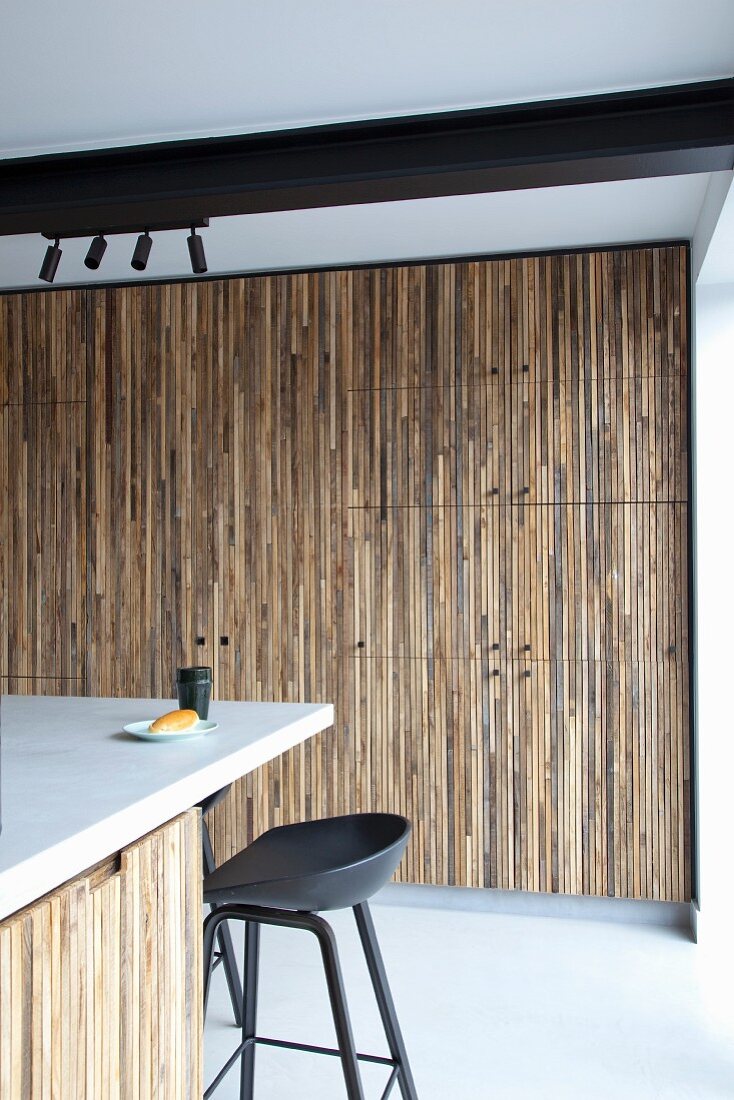 Küchentheke und schwarzer Barhocker vor Einbauschrank mit Holzfront aus schmalen Holzleisten, an Decke schwarzer Stahlträger