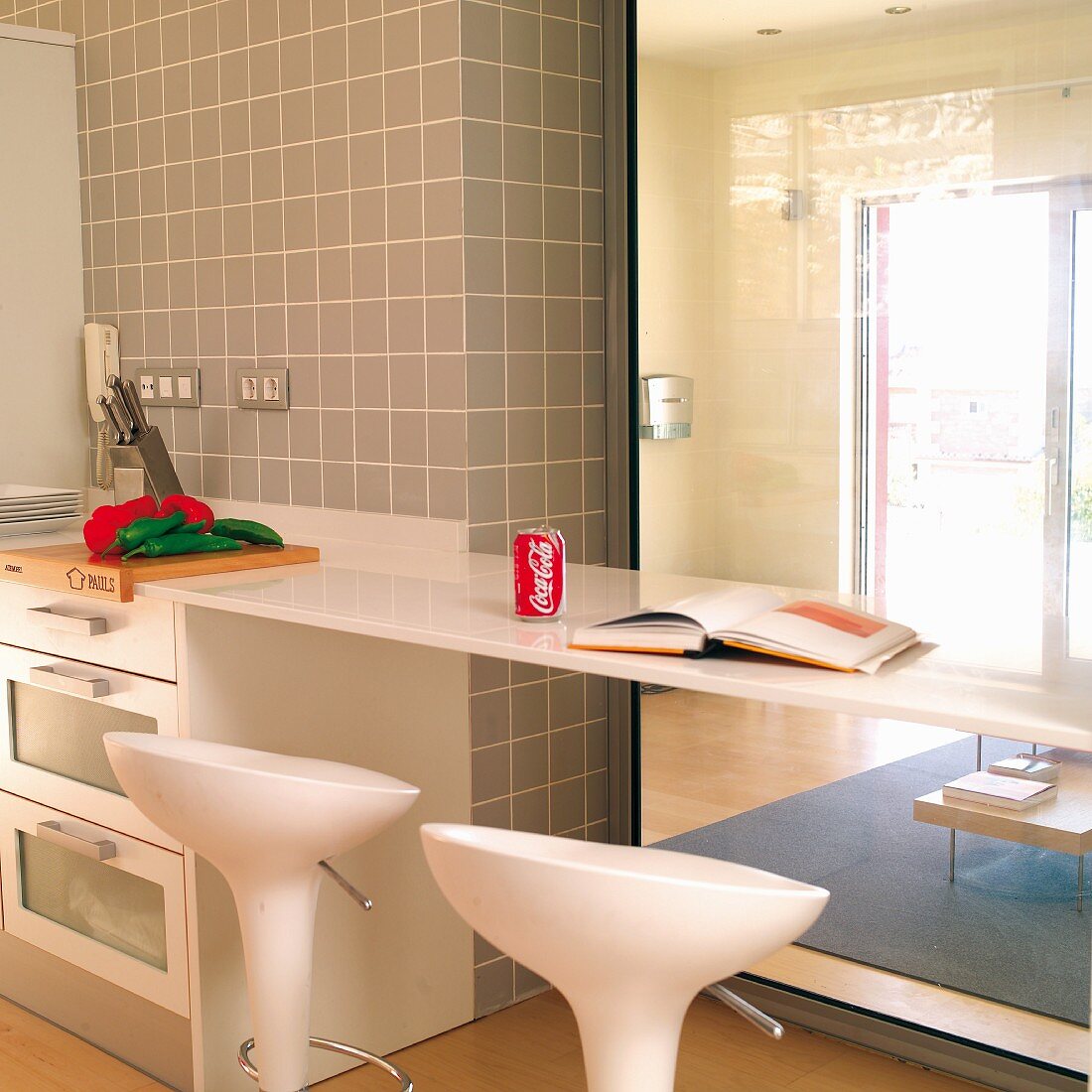 Küchenzeile mit verlängerter Thekenplatte und weiße Barhocker, vor Glasscheibe mit Blick in Wohnzimmer
