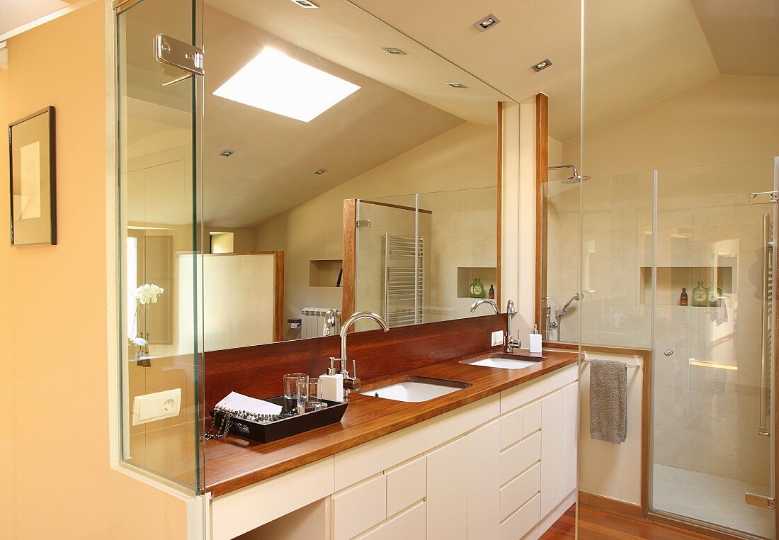 Blick ins Designerbad, seitlich eingebauter Waschtisch mit Unterschränken in Weiß, vollflächiger Spiegel an Wand im Hintergrund Glas Duschkabine