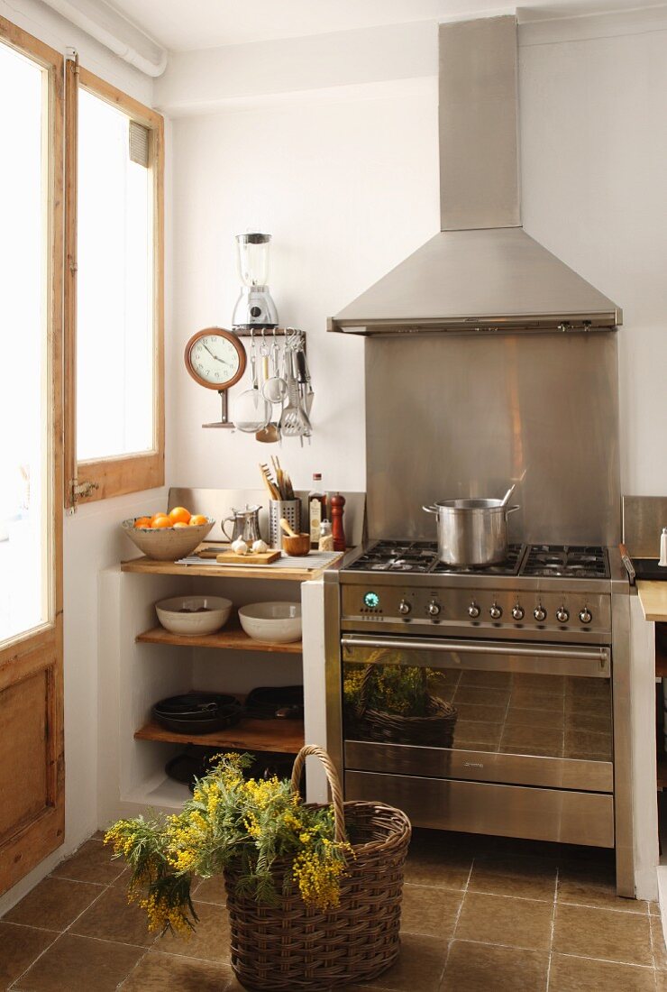 Modern, stainless steel cooker and extractor hood next to shelves of crockery with shopping basket in foreground