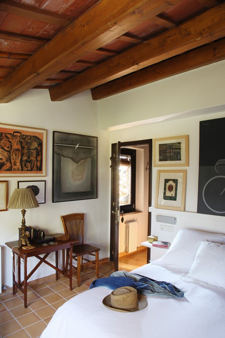 Umlaufend mit Bildern dekoriertes Schlafzimmer mit Holzbalkendecke und Fliesenboden; Beistelltisch und Stuhl im mediterranen Stil