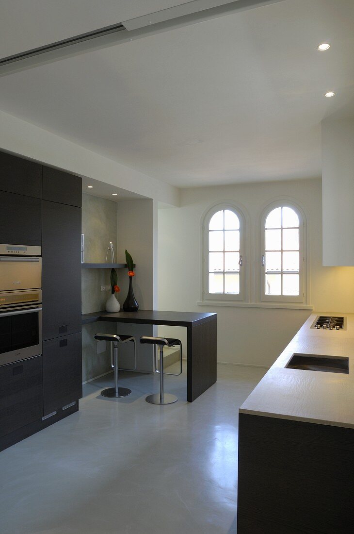 Minimalistische Küchenzeile, gegenüber Einbauschrank mit Küchengeräten und Frühstückstheke mit Barhockern, im Hintergrund Rundbogenfenster