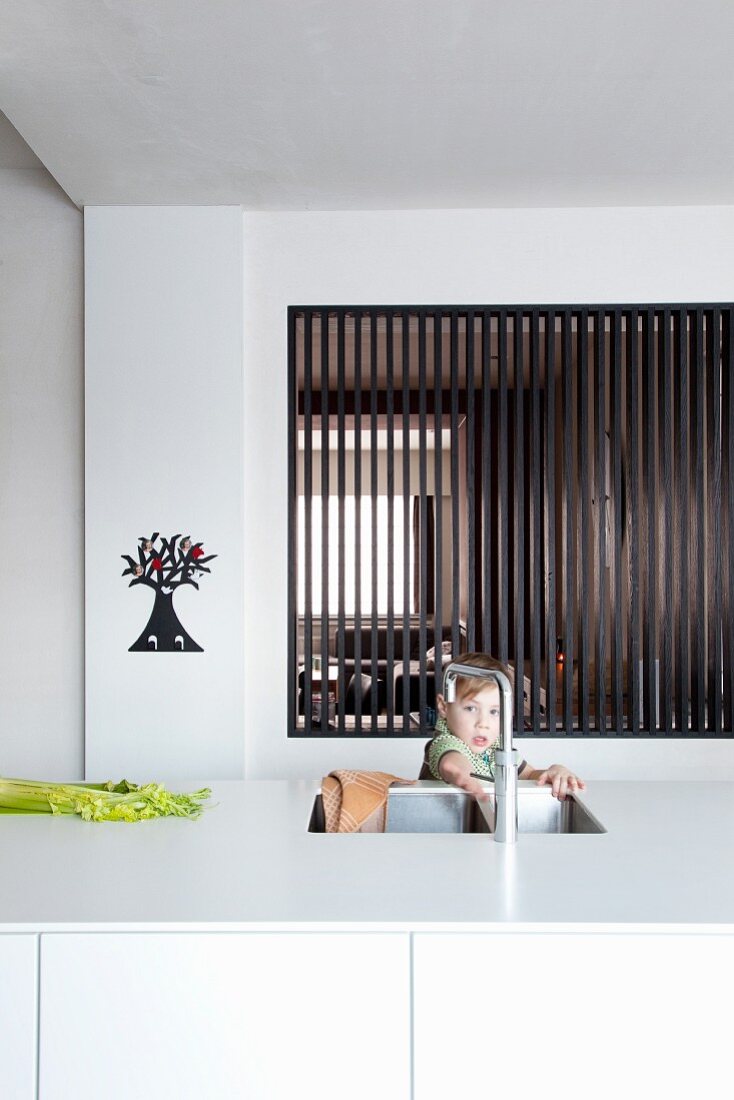 Kind hinter weisser Kücheninsel mit Spülbecken, im Hintergrund Durchreiche mit dunklen Holzlamellen
