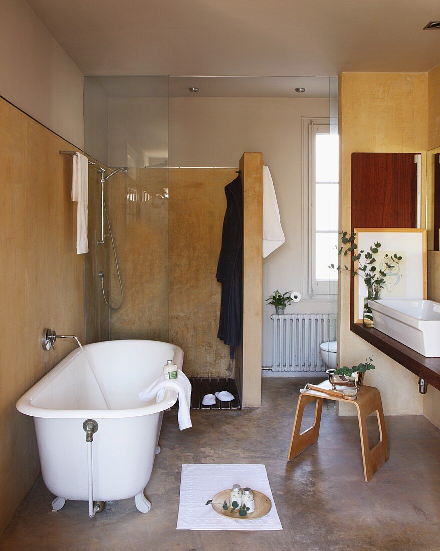 Freistehende Vintage Badewanne vor bodenebener Dusche mit Glastür, seitlich Holzhocker vor Waschtisch in teilweise renoviertem Bad