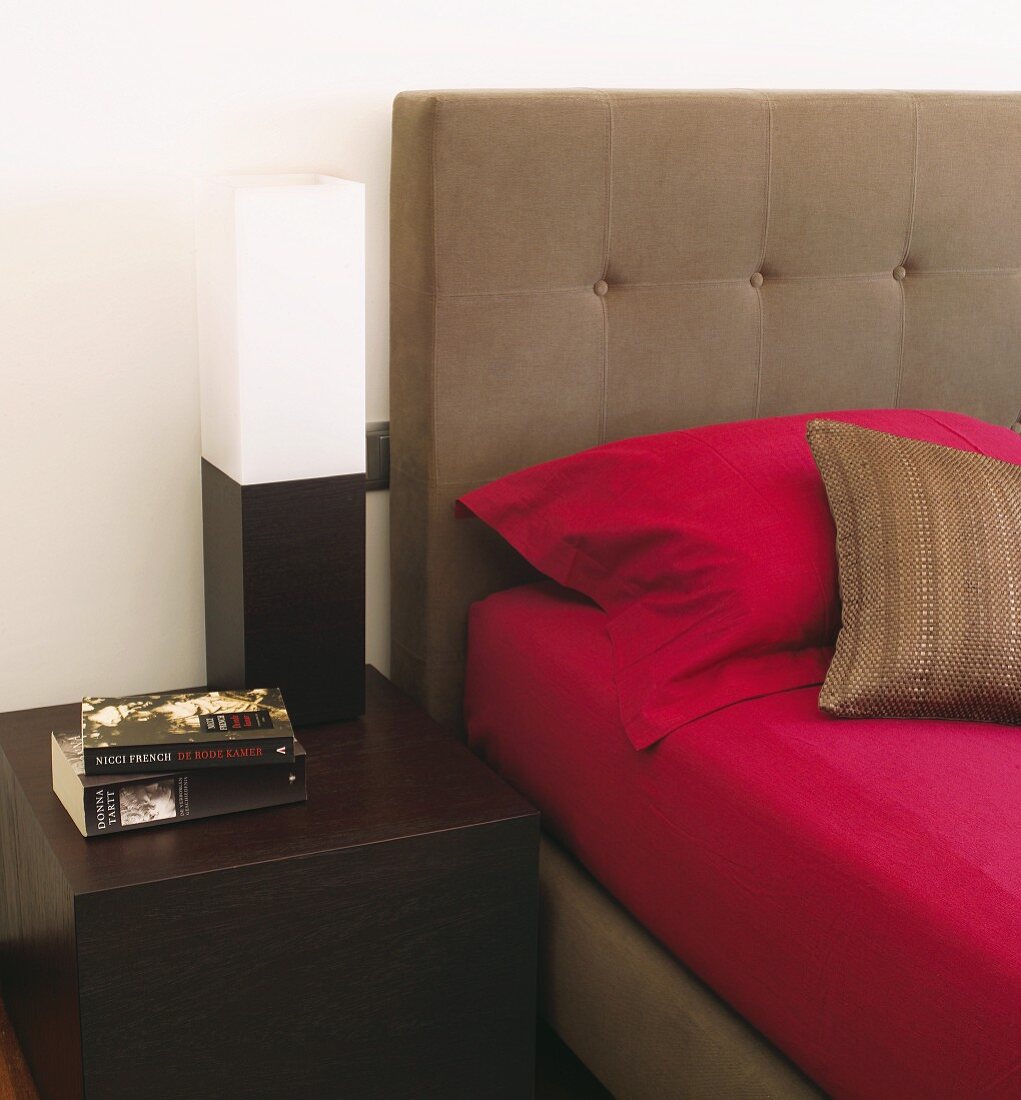 Magentafarbene Bettwäsche auf Polsterbett in warmem Taupe, daneben ein dunkler Nachttischwürfel und kubische Schwarzweiss-Leuchte
