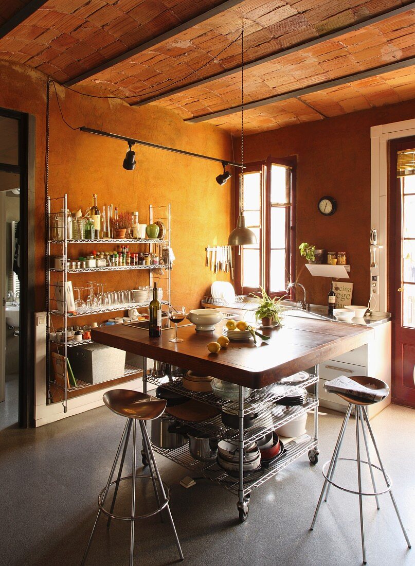 Bar stools with copper seats and mobile breakfast bar in loft-apartment kitchen with brick ceiling; open-fronted metal shelving against wall