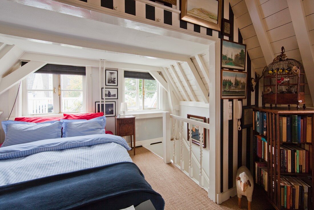 Double bed next to head of staircase in white, wood-clad attic room; wallpaper with wide stripes, ornamental bird cage and bookcase in foreground
