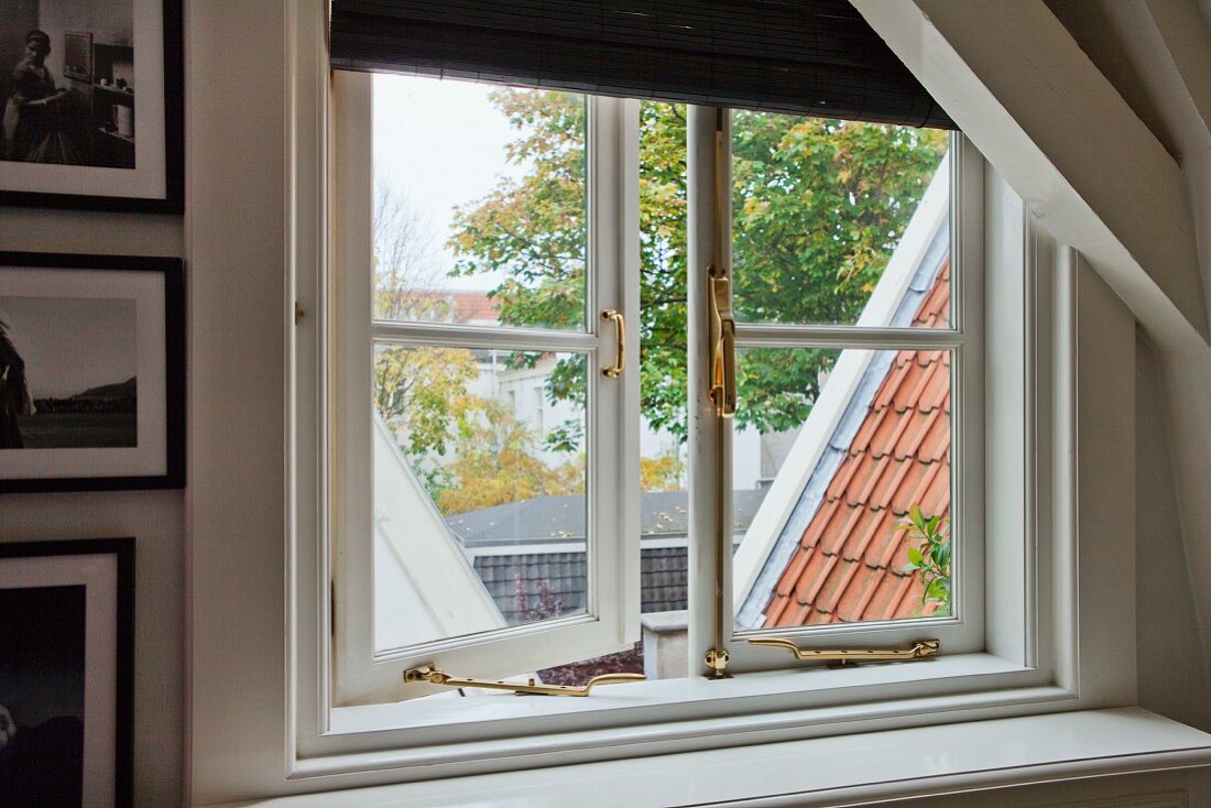 Lattice window with brass fittings in gable wall of attic room; view of roofs and trees
