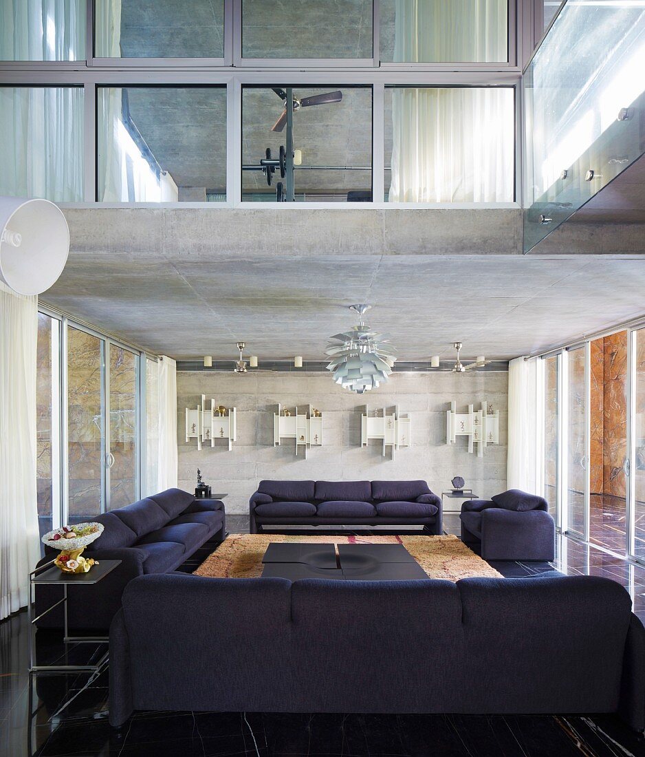 Dunkle Sofagarnitur unter Sichtbetondecke mit Klassiker Hängeleuchte in modernem Wohnraum und Blick auf Zimmer im Obergeschoss