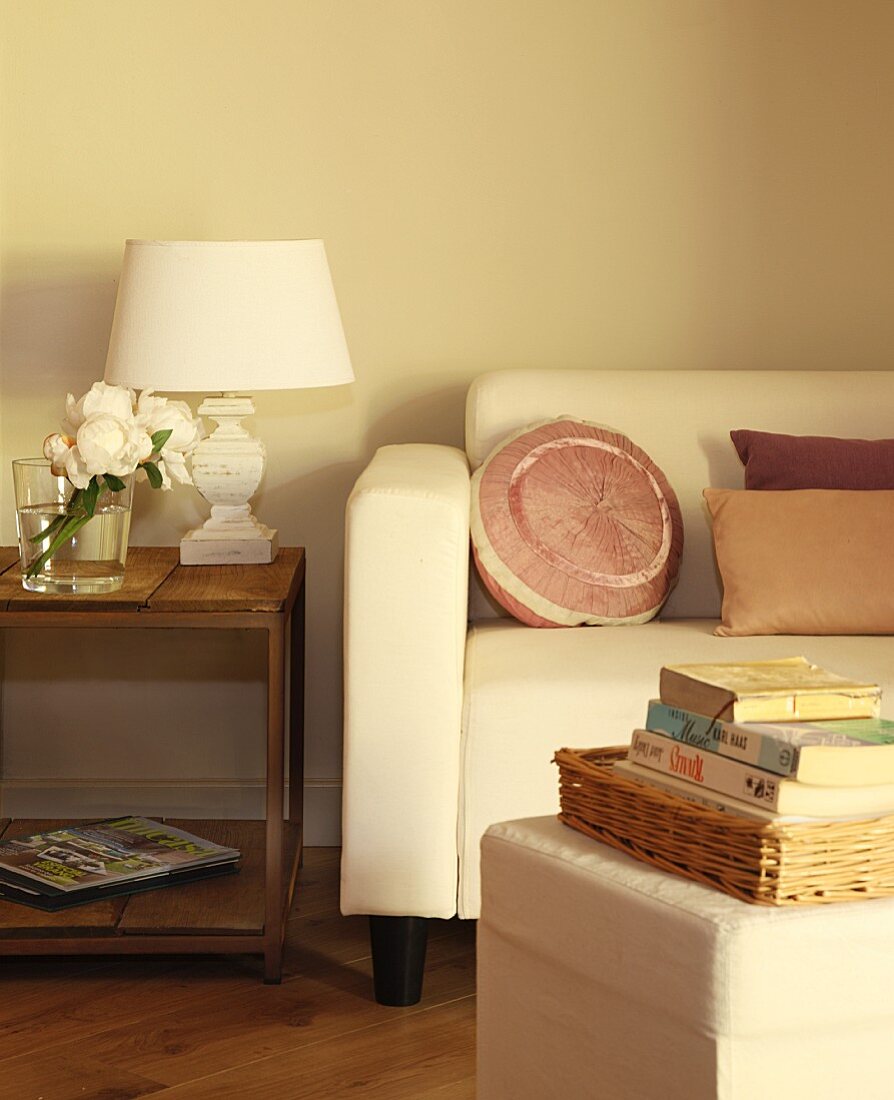 Bücherstapel auf Hocker gegenüber Sofa mit eleganten Kissen, daneben Beistelltisch aus Holz und Tischlampe mit weißem Schirm im Vintage Stil