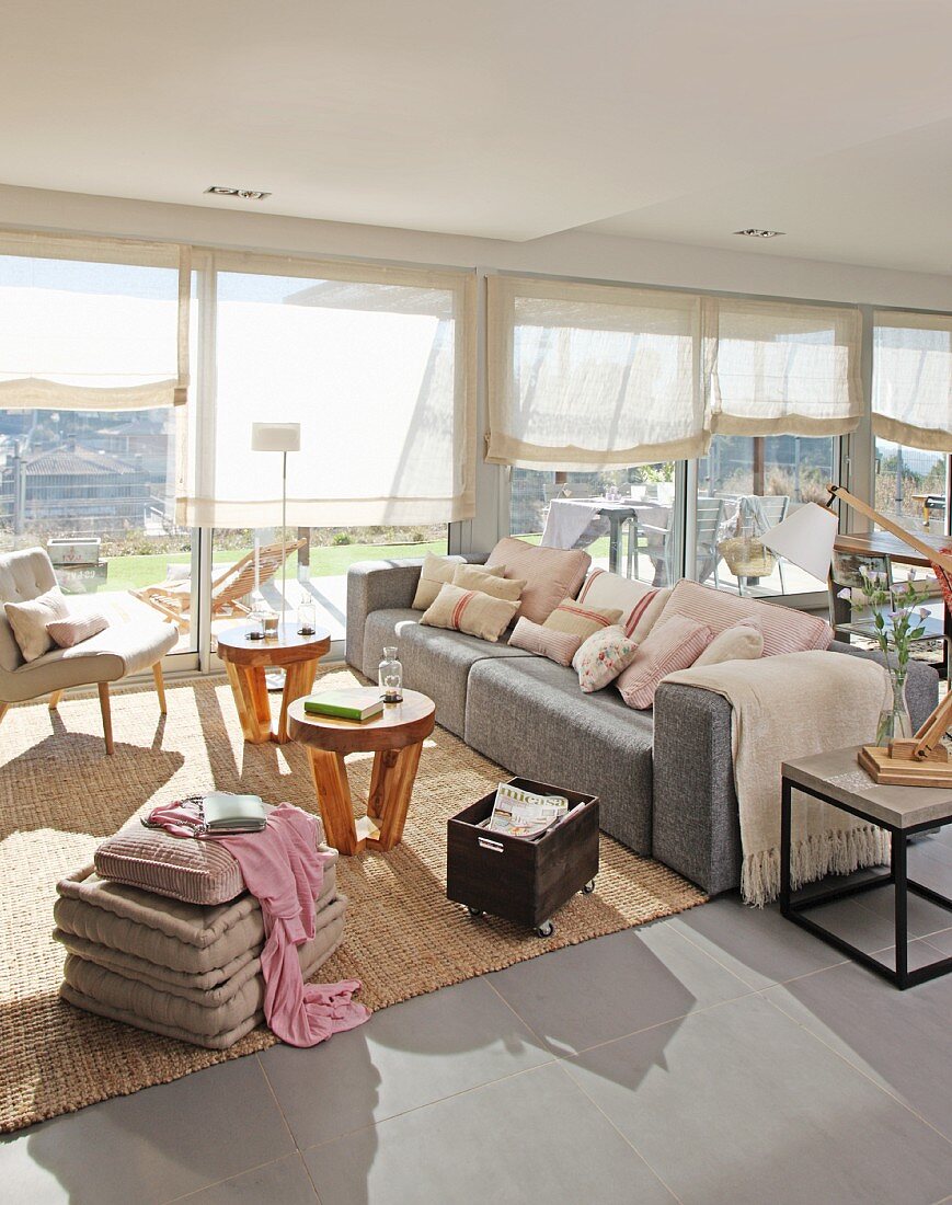 Sonnendurchfluteter Wohnraum mit transparenten Vorhängen am Fenster, im Loungebereich Kissenstapel auf Boden, Polstersessel und hellgraues Sofa um runde Holz Beistelltische