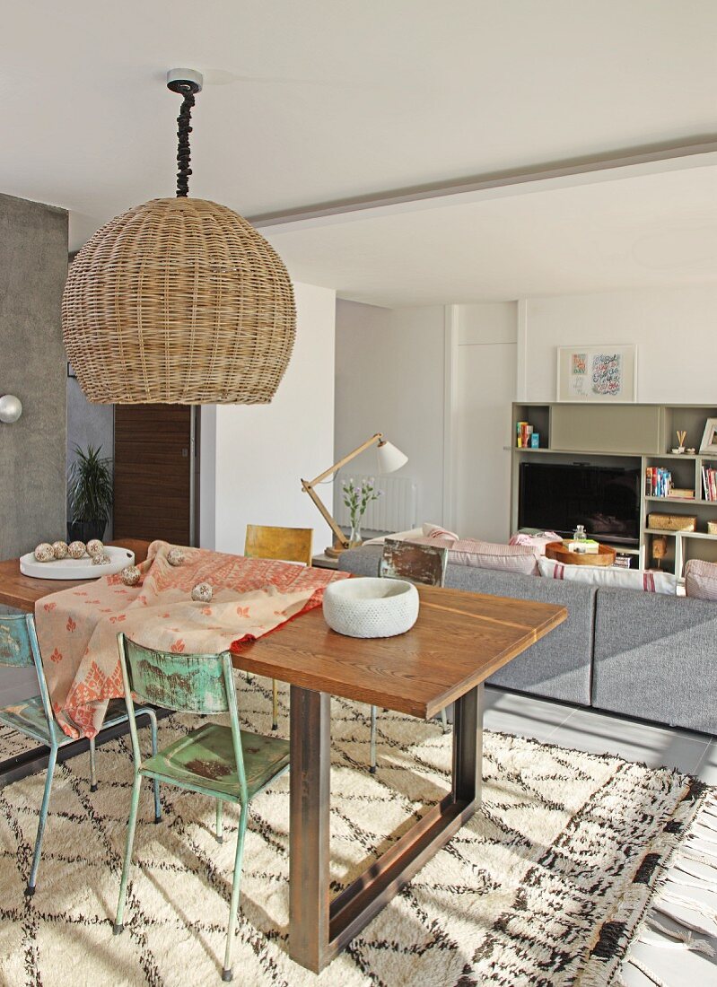 Hängeleuchte mit Rattanschirm über Essplatz mit Vintage Stühlen, auf Boden gemusterter Teppich, im Hintergrund Loungebereich in modernem Wohnraum