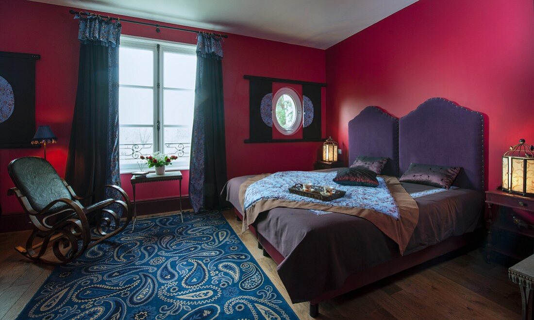 Doppelbett mit violettem Kopfteil, Schaukelstuhl auf Teppich mit Paisleymuster im Schlafzimmer mit roten Wänden