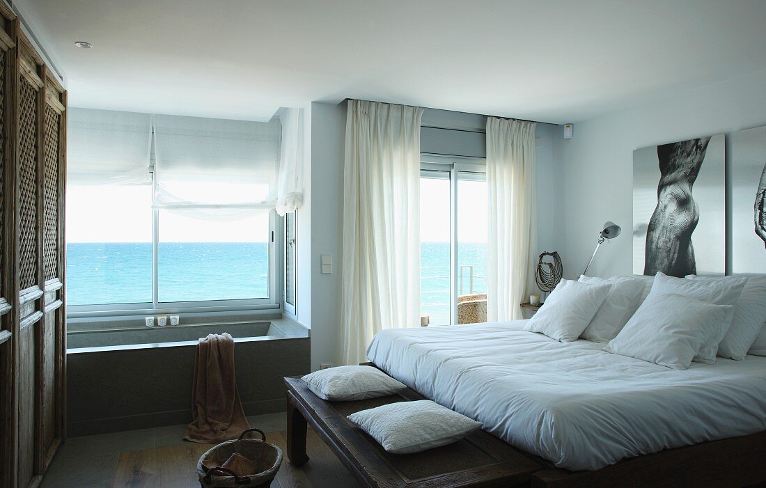 Doppelbett mit weisser Bettwäsche und Badewanne vor dem Fenster mit Panoramablick auf das Meer