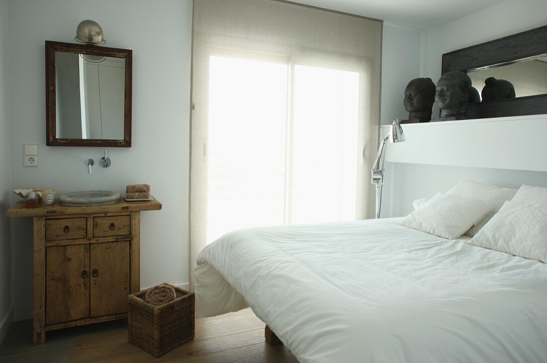 Double bed with white bed linen next to window and rustic chest of drawers with integrated sink