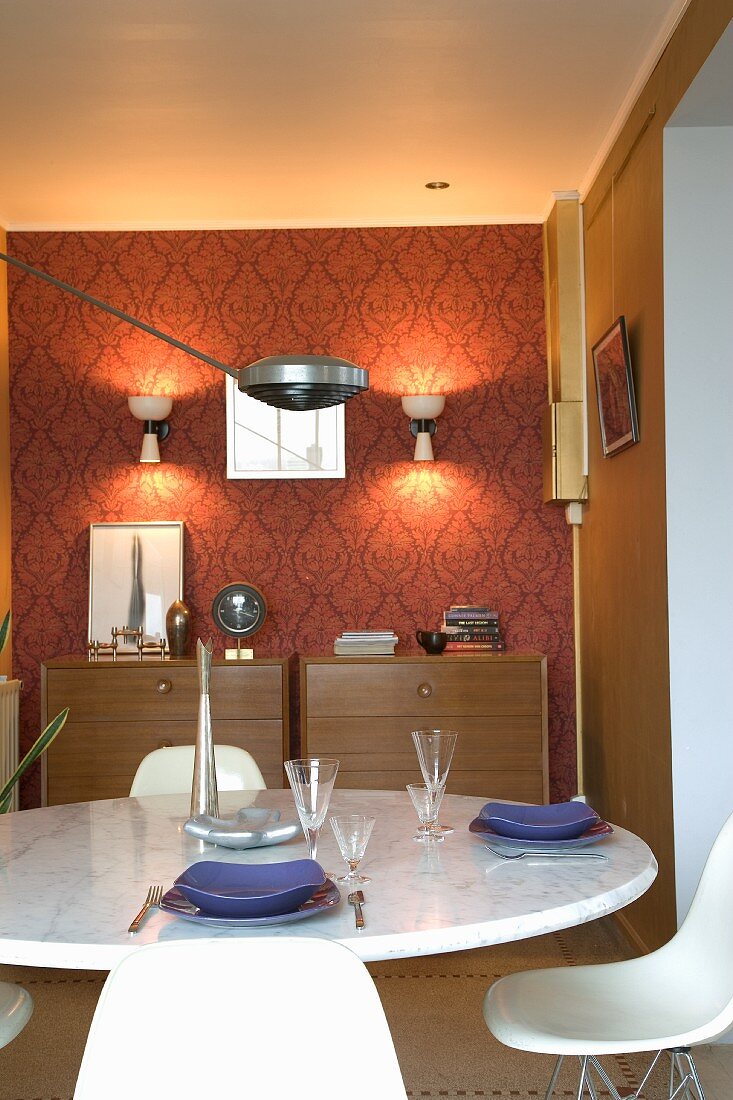 Classic designer furnishings in retro dining room: set table in front of two chests of drawers against red patterned wallpaper